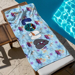 May include: A light blue beach towel featuring a pattern of snowflakes, cartoon characters, and the word "SOON" in a whimsical font. The towel is draped over a wooden lounge chair next to a pool.