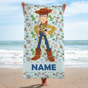 May include: A light blue rectangular beach towel featuring a large central illustration of a cowboy character wearing a brown hat, yellow shirt, and blue pants. The background has a repeating pattern of cartoon characters and the word NAME at the bottom.