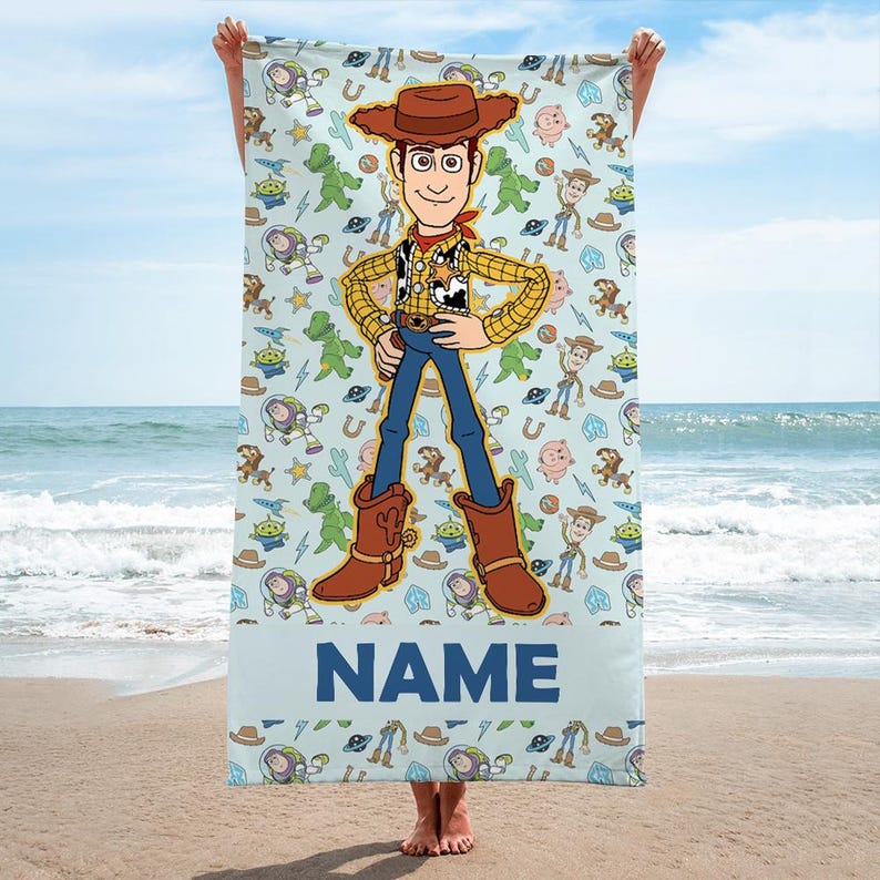 May include: A light blue rectangular beach towel featuring a large central illustration of a cowboy character wearing a brown hat, yellow shirt, and blue pants. The background has a repeating pattern of cartoon characters and the word NAME at the bottom.