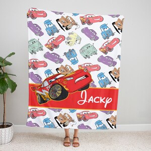 Pu&ograve; includere: Una coperta in pile bianca con un motivo ripetuto di auto dei cartoni animati in rosso, blu, viola e beige. Un'ampia fascia orizzontale rossa al centro mostra il nome Jacky in corsivo bianco, con un grande grafico di un'auto da corsa.