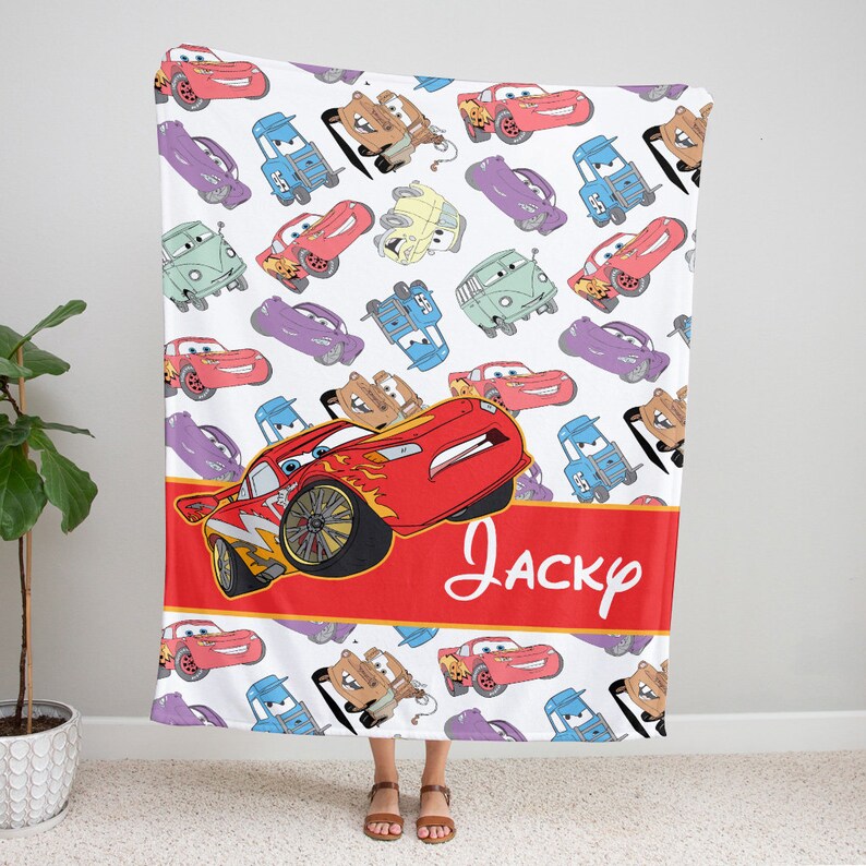 Pu&ograve; includere: Una coperta in pile bianca con un motivo ripetuto di auto dei cartoni animati in rosso, blu, viola e beige. Un'ampia fascia orizzontale rossa al centro mostra il nome Jacky in corsivo bianco, con un grande grafico di un'auto da corsa.