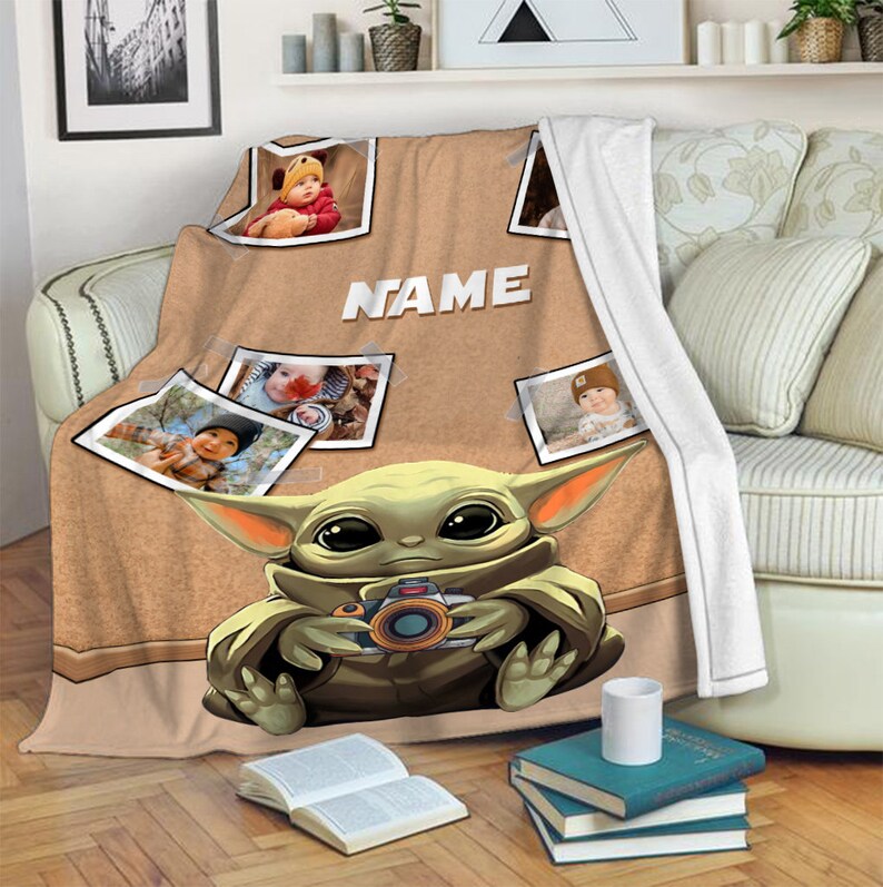 May include: A brown and tan fleece blanket with a cartoon image of a green baby Yoda holding a camera. The blanket has a corkboard background with photos of children and the word "NAME" printed on it.