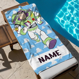 May include: A blue beach towel with a cartoon of Buzz Lightyear running. The towel has a light blue sky background with white clouds and a white stripe at the bottom with the word "NAME" in dark purple.