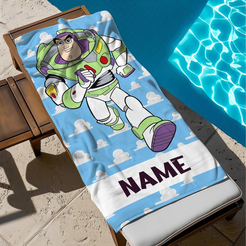 May include: A blue beach towel with a cartoon of Buzz Lightyear running. The towel has a light blue sky background with white clouds and a white stripe at the bottom with the word "NAME" in dark purple.