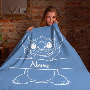 May include: A light blue fleece blanket featuring a large white line-art illustration of the character Stitch from Lilo & Stitch. The design includes a horizontal white banner across the center with the word Name printed in white text.