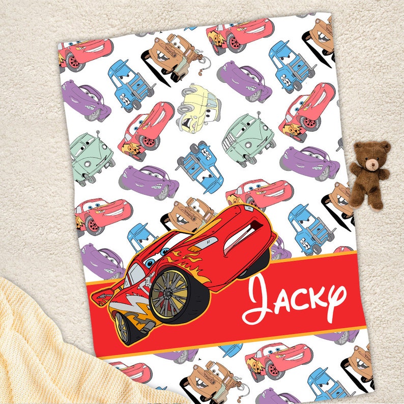 Pu&ograve; includere: Una coperta bianca con un motivo ripetuto di colorate auto dei cartoni animati del film Cars, inclusi veicoli rossi, blu, viola e marroni. Una grande auto rossa &egrave; centrata in basso con il nome Jacky scritto in corsivo bianco.