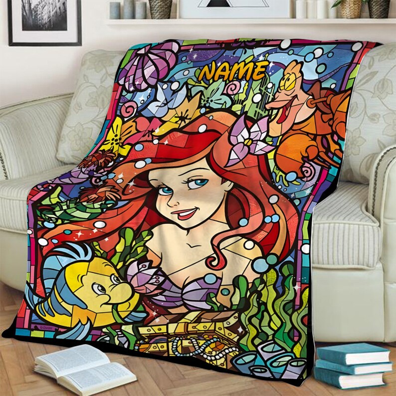 Puede incluir: Una manta rectangular con una colorida ilustraci&oacute;n estilo vidriera que muestra a una sirena de pelo rojo, un pez amarillo y azul, un cangrejo y un cofre del tesoro. La palabra NAME est&aacute; impresa en amarillo en la parte superior.