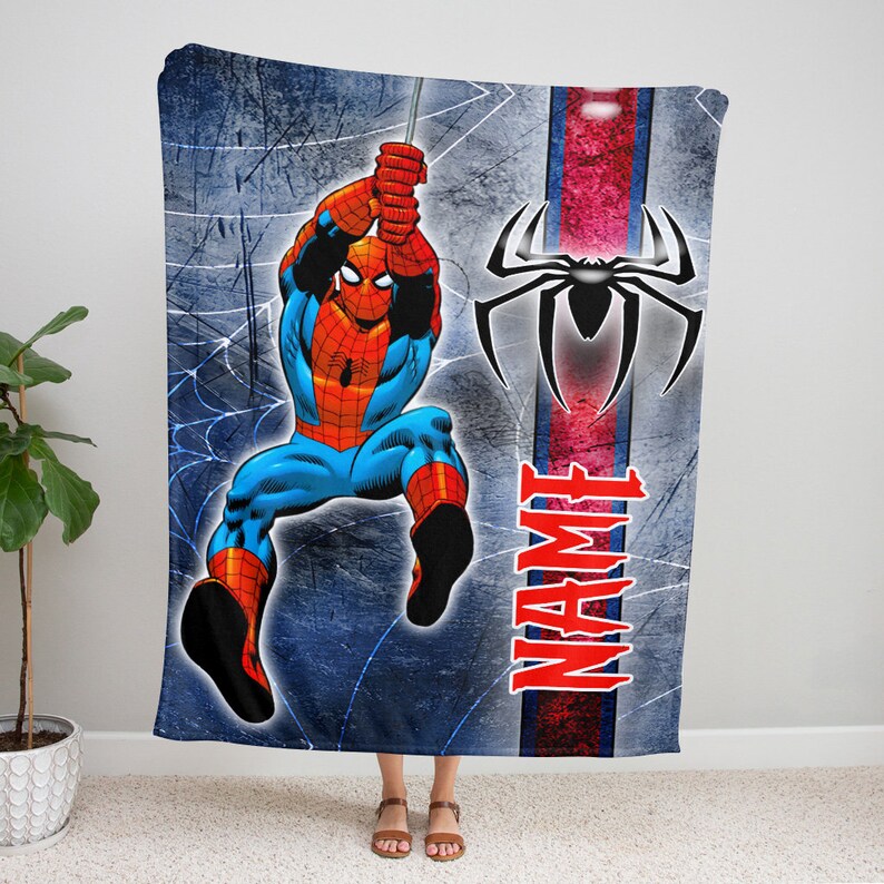 以下が含まれることがあります： クモの巣からぶら下がるスパイダーマンのグラフィックが描かれた長方形のフリースブランケット。背景は質感のある青色で、黒いクモのエンブレムと赤い大文字でNAMEと書かれた縦の赤いストライプが入っています。