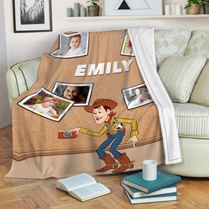 May include: A brown and tan fleece blanket featuring a cartoon image of Woody from Toy Story taking a photograph. The blanket has a corkboard background with photos of children and the name "EMILY" written in white letters.