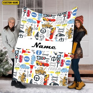 May include: A white fleece blanket featuring a repeating pattern of cartoon race cars, tow trucks, traffic lights, wrenches, and road signs. A horizontal white band across the centre displays the word Name in black script text.