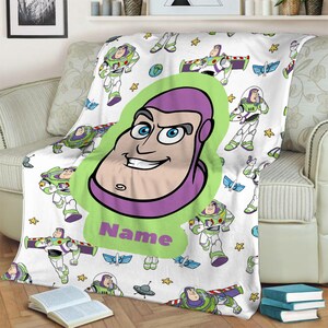 May include: A white fleece blanket featuring a repeating pattern of Buzz Lightyear in various poses, small yellow stars, and blue wings. A large central lime green shape displays a cartoon portrait of Buzz Lightyear and the word Name.