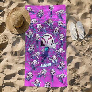 K&ouml;nnte beinhalten: Ein rechteckiges Strandtuch mit einem leuchtend pinkfarbenen Hintergrund, das ein wiederkehrendes Muster von Ghost-Spider-Figuren in verschiedenen Posen zeigt, &uuml;berlagert mit einem blauen Spinnennetz-Design und dem Wort NAME in blauen Buchstaben.