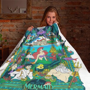 May include: A white throw blanket featuring a colorful graphic print of The Little Mermaid characters including Ariel, King Triton, Prince Eric, and Ursula. The design displays an underwater scene with coral, sea life, and a castle, with the text The Little Mermaid at the bottom.