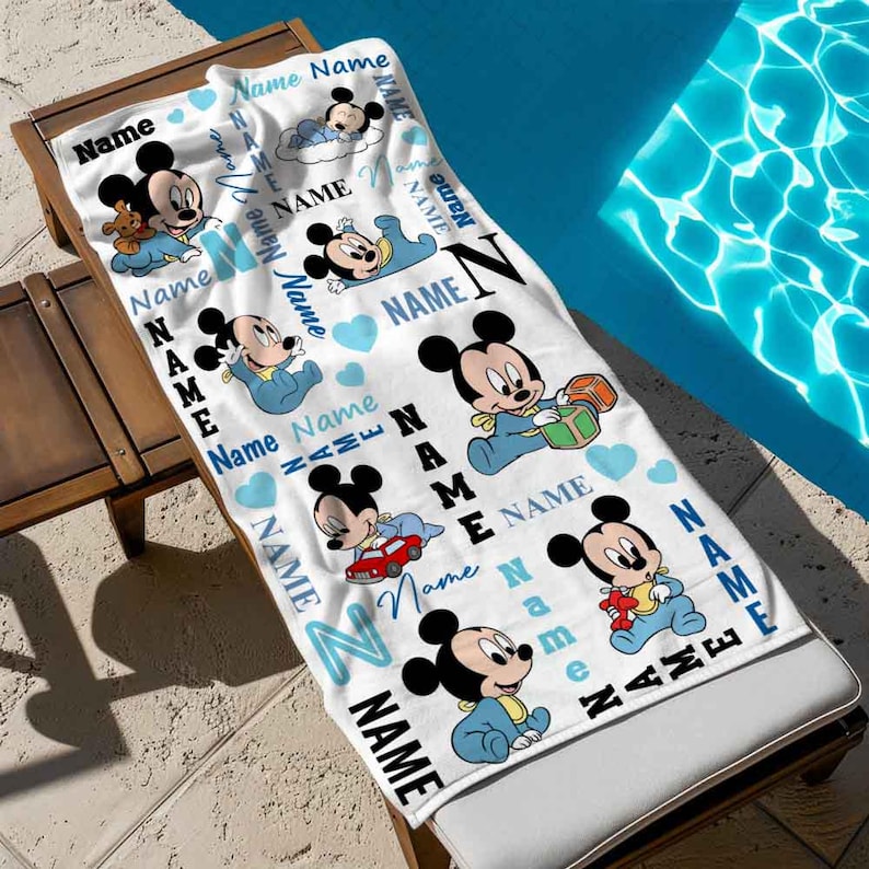 May include: White beach towel with a blue and white pattern featuring Mickey Mouse characters. The towel has the word "Name" repeated throughout the design.