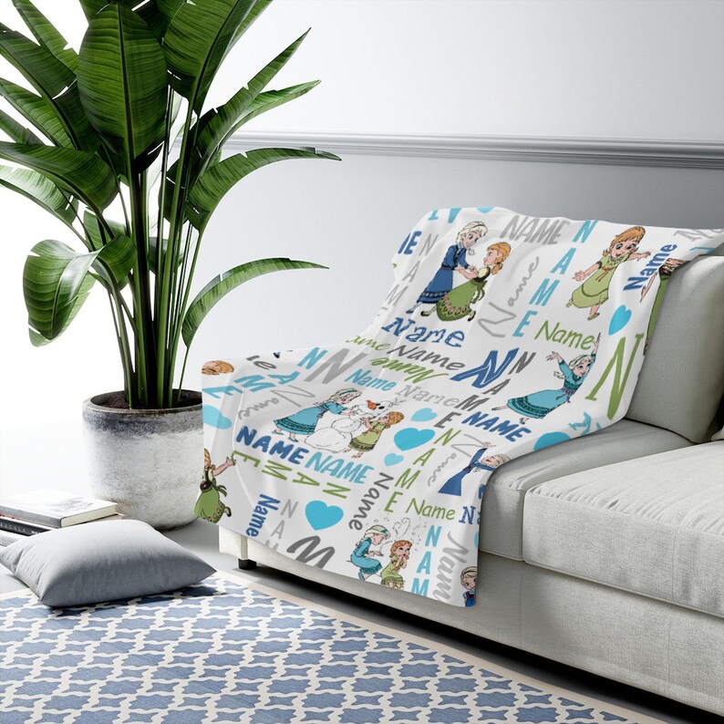 May include: A white throw blanket featuring a repeating pattern of two cartoon characters in blue and green dresses, a snowman, blue hearts, and the word Name in various blue, green, and grey fonts, arranged in a scattered, multi-directional layout.