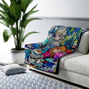 Pode incluir: Uma manta macia com um gr&aacute;fico colorido em estilo vitral que mostra Elsa, Anna e Olaf de Frozen. O design geom&eacute;trico em tons de azul, roxo e rosa inclui a palavra NAME impressa em azul claro na parte inferior da pe&ccedil;a.