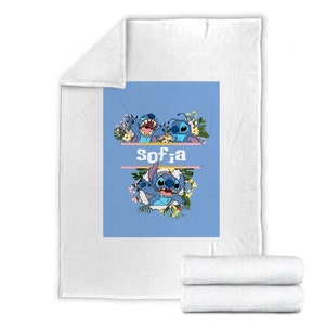 May include: A white fleece blanket featuring a central light blue rectangular graphic. The design displays the blue character Stitch surrounded by tropical flowers and leaves, with the name Sofia printed in white letters between two horizontal lines.