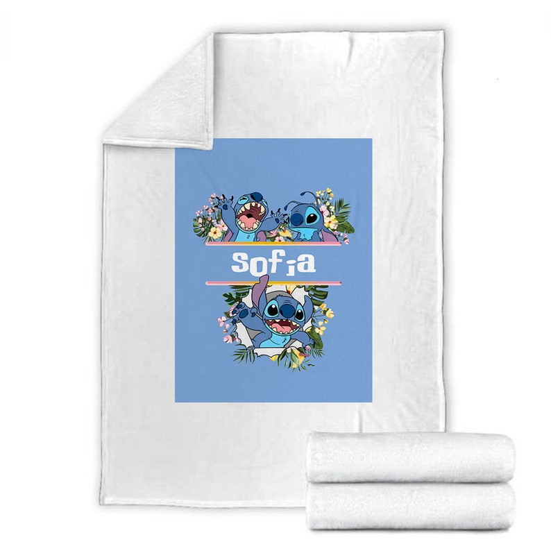 May include: A white fleece blanket featuring a central light blue rectangular graphic. The design displays the blue character Stitch surrounded by tropical flowers and leaves, with the name Sofia printed in white letters between two horizontal lines.