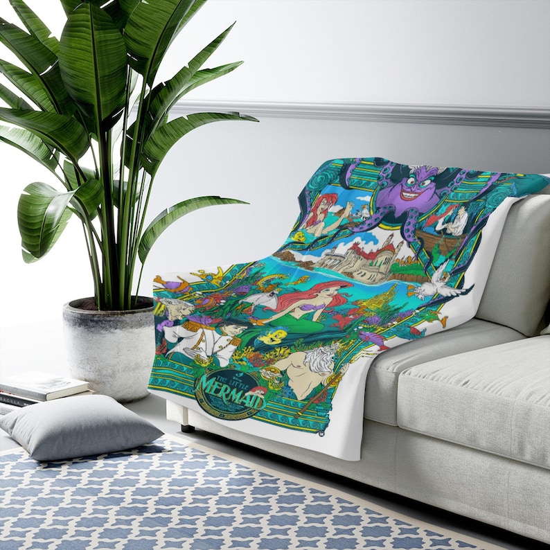 May include: A white throw blanket featuring a colorful graphic print of The Little Mermaid characters including Ariel, King Triton, Prince Eric, and Ursula. The design incorporates underwater scenes, a castle, and the text The Little Mermaid.