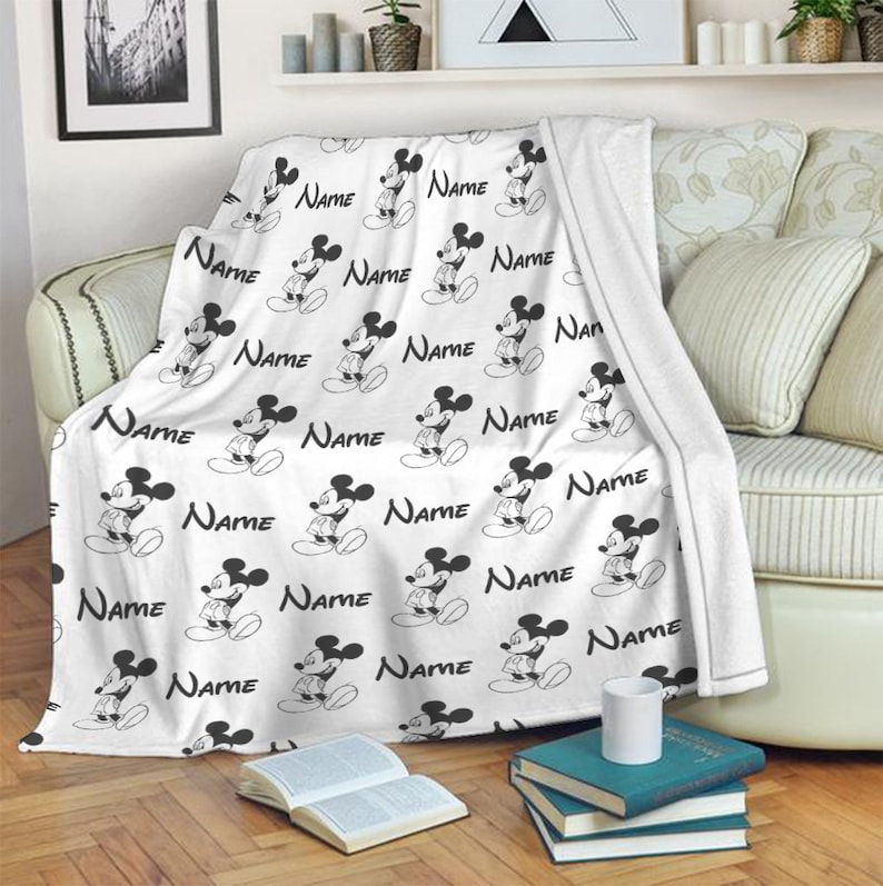 May include: A white fleece throw blanket featuring a repeating black and white pattern of Mickey Mouse figures and the word Name printed in a script font. The blanket is draped over a sofa, showcasing the soft texture and the character design.