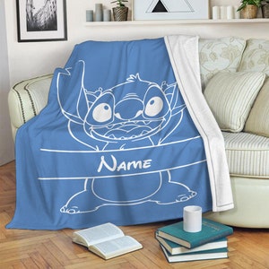 May include: A blue fleece throw blanket featuring a white line art illustration of the character Stitch from Lilo and Stitch. The design includes a horizontal white banner across the center with the word Name printed in white capital letters.
