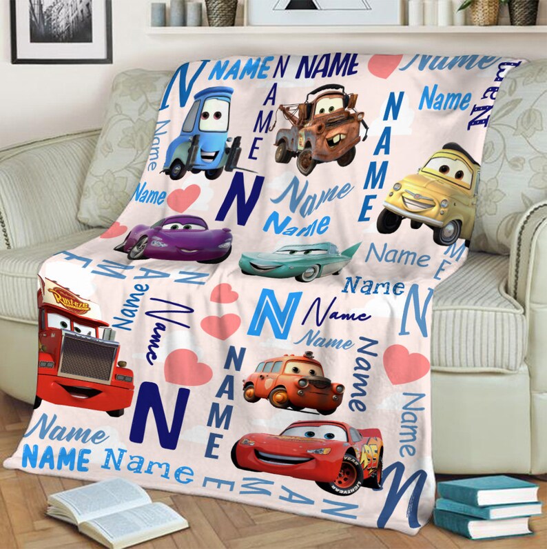 May include: A soft fleece blanket featuring a repeating pattern of various cartoon cars from the movie Cars, including Lightning McQueen, Mater, and others. The design includes the word Name in blue text, the letter N, and pink hearts.