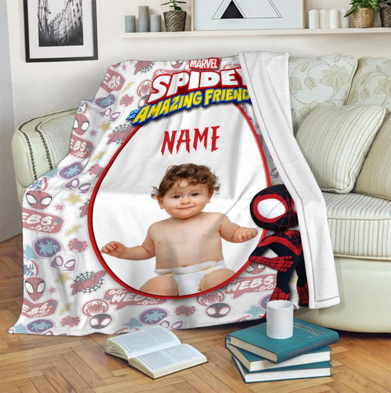 May include: A white fleece blanket featuring a Marvel Spidey and His Amazing Friends logo, a red circular frame for a custom photo, the text NAME, and a repeating pattern of spider-themed icons, stars, and text bubbles with a plush toy attached.