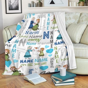 May include: A white fleece throw blanket featuring a repeating pattern of two cartoon characters, one with blonde hair in a blue dress and one with auburn hair in a green dress, building a snowman. The design includes blue hearts and the word Name.