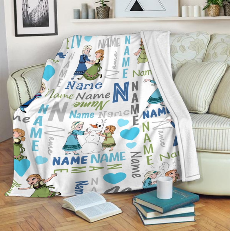 May include: A white fleece throw blanket featuring a repeating pattern of two cartoon characters, one with blonde hair in a blue dress and one with auburn hair in a green dress, building a snowman. The design includes blue hearts and the word Name.