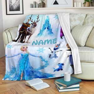 May include: A blue and white fleece blanket with a watercolor design featuring characters from the movie Frozen. The blanket features Elsa, Anna, Olaf, Kristoff, and Sven. The blanket also includes the text "NAME" in blue.