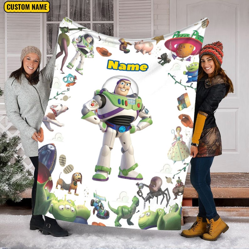 Puede incluir: Una manta de forro polar blanca con un gr&aacute;fico central grande de Buzz Lightyear rodeado de varias ilustraciones de personajes de Toy Story, incluyendo a Woody, Rex y Slinky Dog, con el texto "Name" impreso en azul y amarillo arriba.