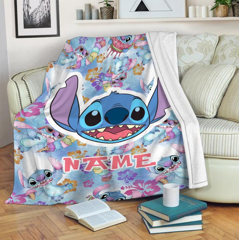 Puede incluir: Una manta de forro polar suave con un patr&oacute;n repetitivo del personaje azul Stitch, flores de hibisco rosas y conos de helado. Un gr&aacute;fico central grande muestra la cara de Stitch, con la palabra NAME impresa en letras de bloque rosas.