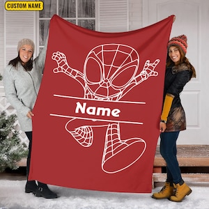 May include: A red fleece blanket featuring a white line art illustration of a cartoon superhero character in a dynamic pose. A horizontal white banner across the center contains the text Name in white letters. The blanket has a solid red background.