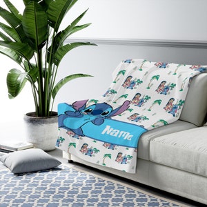 K&ouml;nnte beinhalten: Eine wei&szlig;e Wohndecke mit einem sich wiederholenden Muster aus der blauen Figur Stitch und Palmen. Ein breites blaues horizontales Band in der Mitte zeigt das Wort Name in wei&szlig;er Schrift, dar&uuml;ber befindet sich eine gro&szlig;e Abbildung von Stitch.