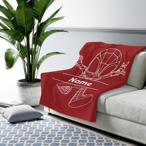 May include: A deep red throw blanket featuring a white line-art graphic of a superhero character in a dynamic pose. The design includes a horizontal line with the word Name printed in white, allowing for personalization on the soft fabric surface.