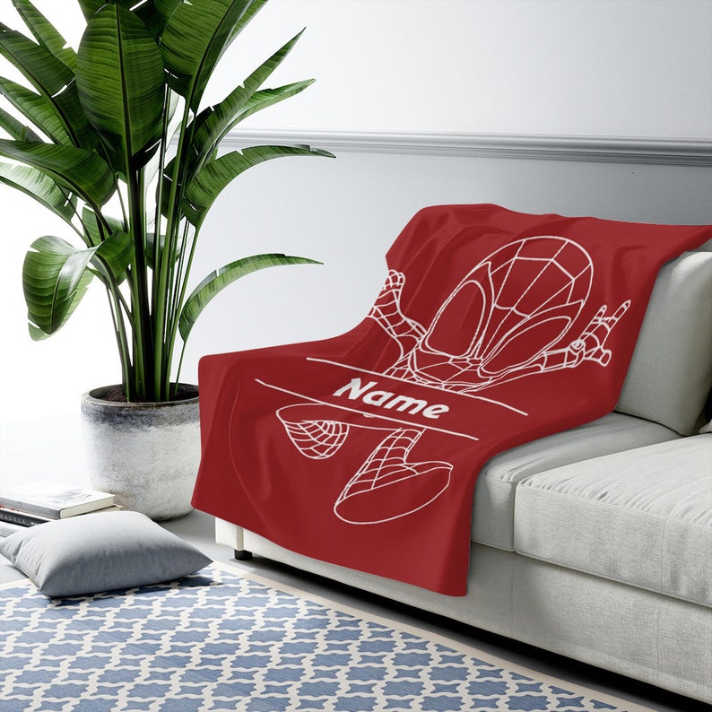May include: A deep red throw blanket featuring a white line-art graphic of a superhero character in a dynamic pose. The design includes a horizontal line with the word Name printed in white, allowing for personalization on the soft fabric surface.