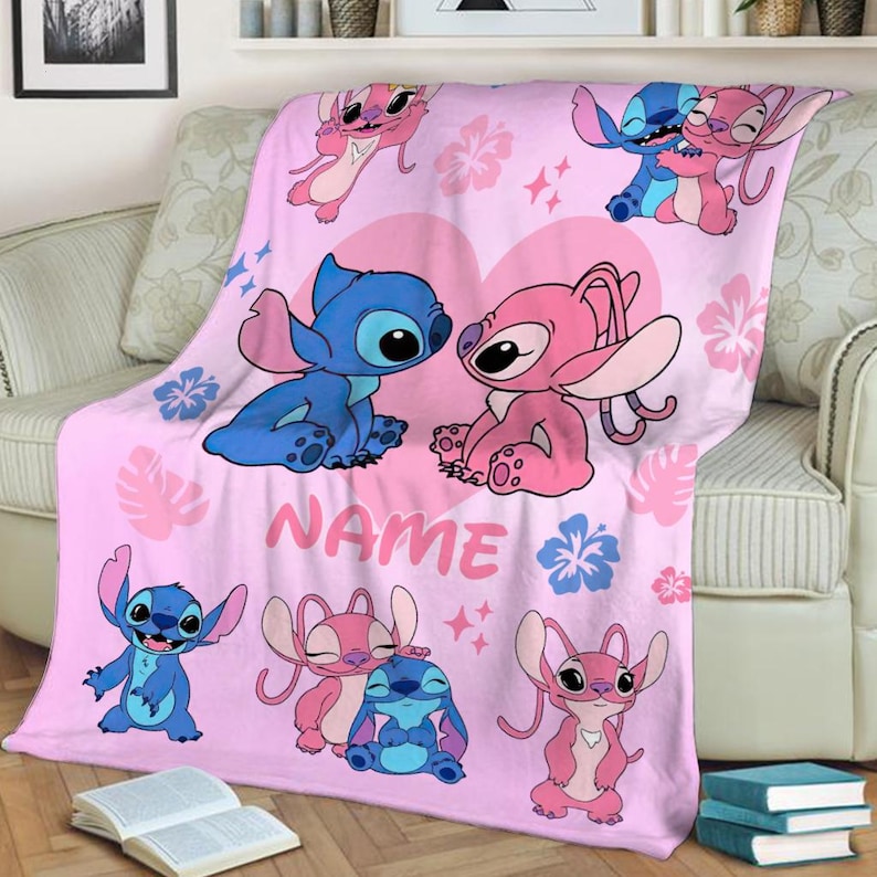 May include: A pink throw blanket featuring cartoon characters in blue and pink, with a large heart design. The blanket has a repeating pattern of the characters and floral accents. The word "NAME" is printed on the blanket.