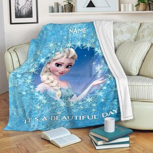 Pu&ograve; includere: Una coperta in pile blu con la grafica di un personaggio biondo con acconciatura a treccia, circondato da un motivo circolare di fiocchi di neve bianchi. Il testo NAME &egrave; stampato in alto e IT'S A BEAUTIFUL DAY &egrave; stampato in basso.