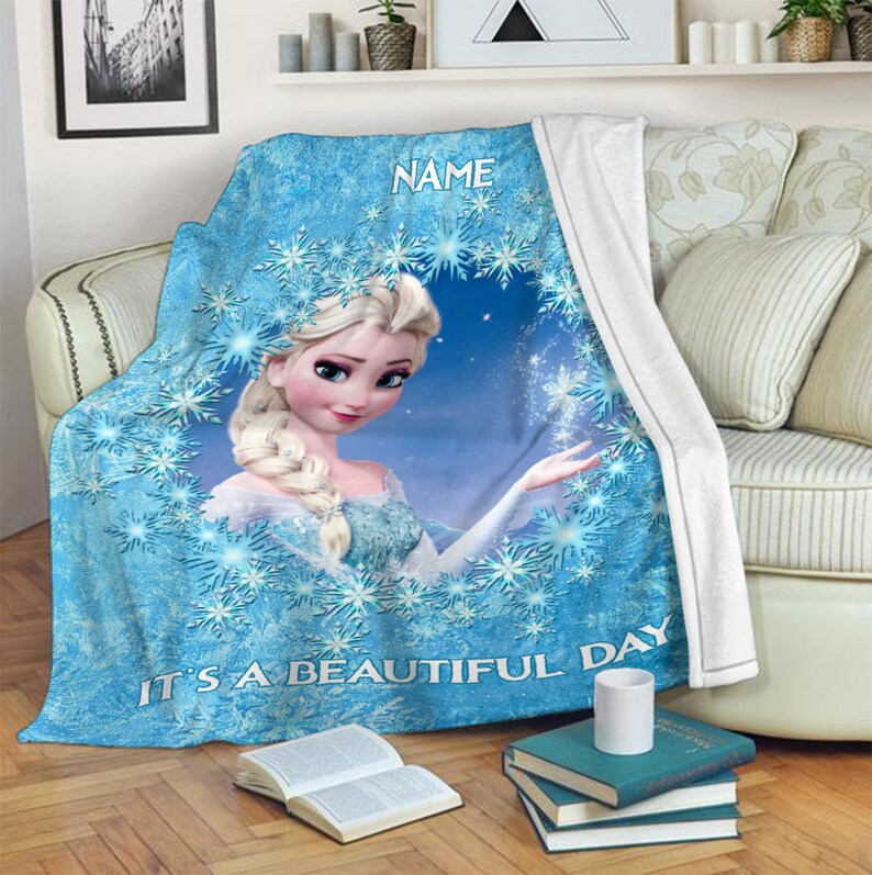 Pu&ograve; includere: Una coperta in pile blu con la grafica di un personaggio biondo con acconciatura a treccia, circondato da un motivo circolare di fiocchi di neve bianchi. Il testo NAME &egrave; stampato in alto e IT'S A BEAUTIFUL DAY &egrave; stampato in basso.