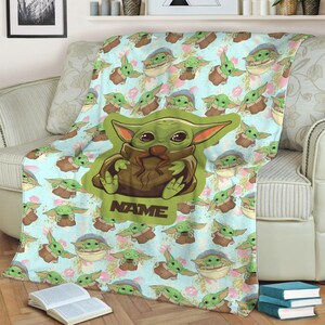 Może przedstawiać: Miękki, niebieski i zielony koc polarowy z powtarzającym się wzorem postaci z kresk&oacute;wek. Postać jest zielona z dużymi oczami i trzyma brązowy przedmiot. Koc ma nadrukowane słowo "NAME".