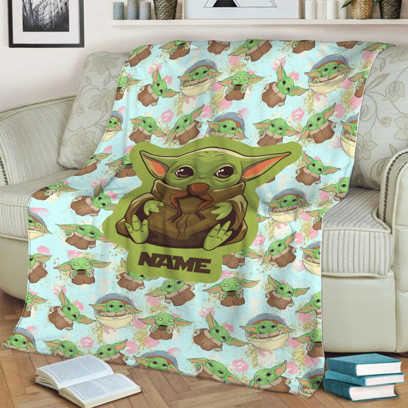 Może przedstawiać: Miękki, niebieski i zielony koc polarowy z powtarzającym się wzorem postaci z kresk&oacute;wek. Postać jest zielona z dużymi oczami i trzyma brązowy przedmiot. Koc ma nadrukowane słowo "NAME".