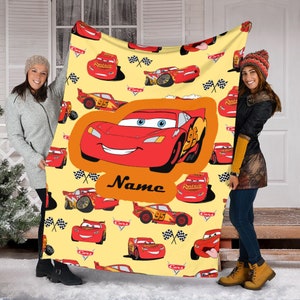 Może przedstawiać: Ż&oacute;łty koc polarowy z powtarzającym się wzorem czerwonych samochod&oacute;w wyścigowych Lightning McQueen z filmu Cars, flagami w szachownicę i logo Cars. Duża pomarańczowa grafika pośrodku przedstawia samoch&oacute;d wyścigowy i słowo Name.