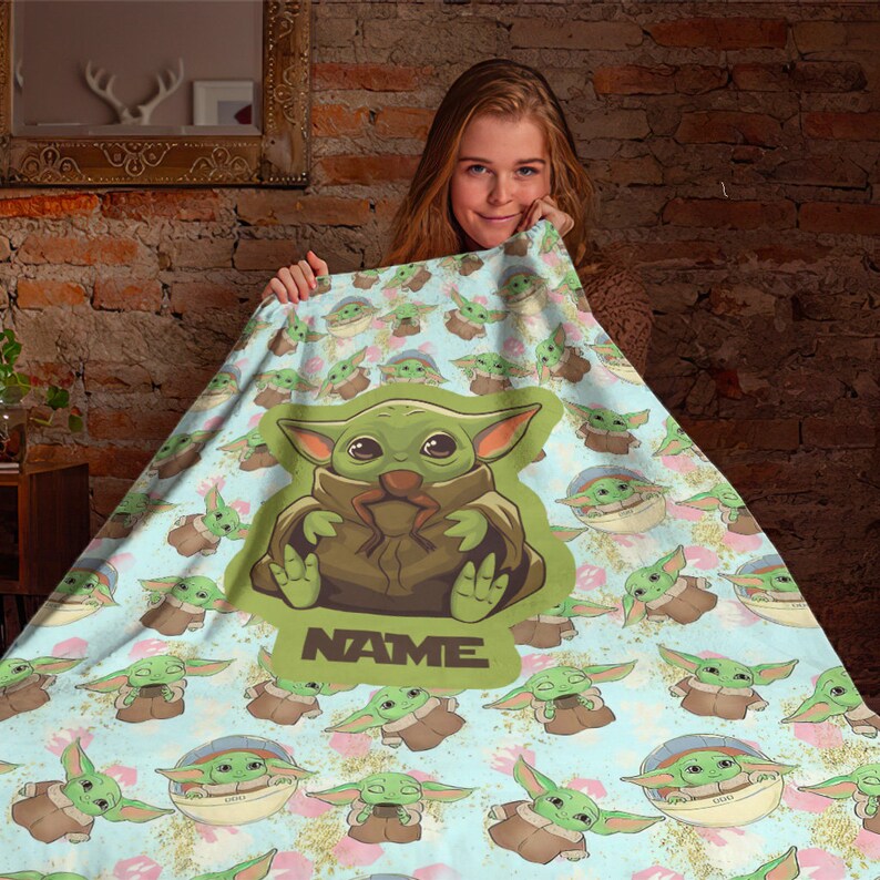 Może przedstawiać: Miękki, niebieski i zielony koc polarowy z powtarzającym się wzorem postaci z kresk&oacute;wek. Postać jest zielona z dużymi uszami i trzyma małą żabę. Koc ma napis "NAME" nadrukowany w środku.