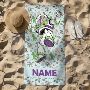 May include: A rectangular beach towel featuring a repeating pattern of Toy Story characters including Buzz Lightyear, Woody, and Rex on a light blue background. A large Buzz Lightyear graphic is centered, and the word NAME is printed in purple.