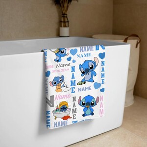 K&ouml;nnte beinhalten: Ein wei&szlig;es Handtuch mit einem sich wiederholenden Muster der blauen Figur Stitch in verschiedenen Posen, darunter in einer Badewanne sitzend, ein Spielzeug haltend und mit einem Auto spielend. Das Design enth&auml;lt blaue Herzen und das Wort NAME in blauem und rosa Text.
