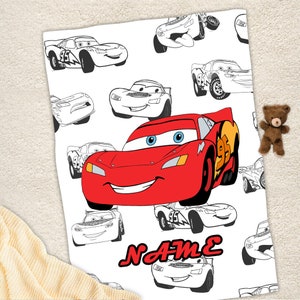 May include: A white blanket featuring a repeating pattern of black and white line art of race cars, with one large, central, full-color red race car character with blue eyes and the number 95 on its side. The word NAME is printed in red.