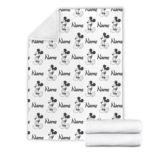 May include: A white fleece blanket featuring a repeating black and white pattern of Mickey Mouse figures and the word Name printed in a script font. The blanket is shown unfolded with a folded section at the bottom and a corner folded over.