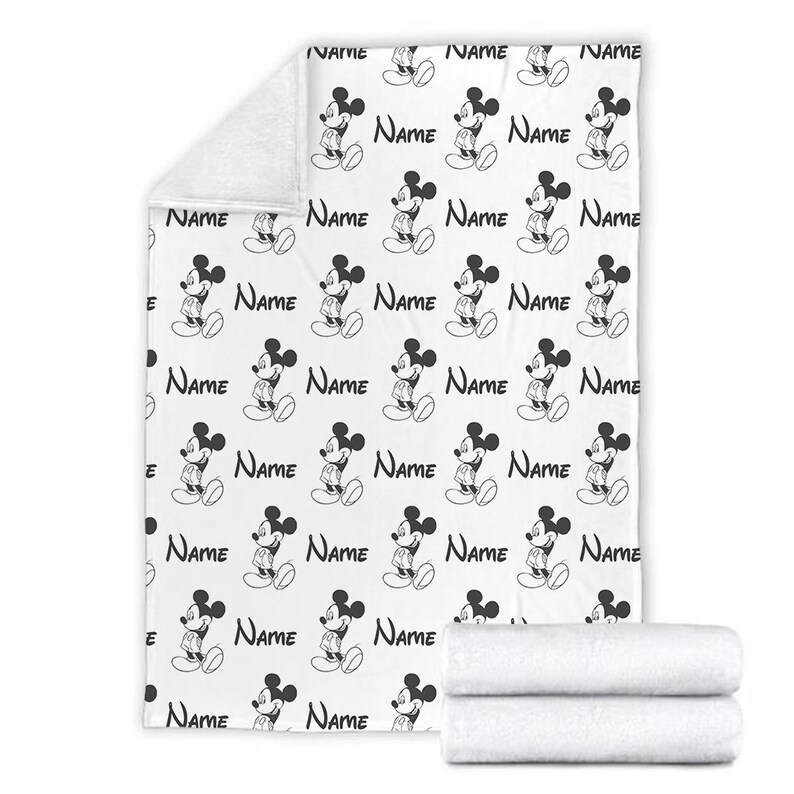 May include: A white fleece blanket featuring a repeating black and white pattern of Mickey Mouse figures and the word Name printed in a script font. The blanket is shown unfolded with a folded section at the bottom and a corner folded over.