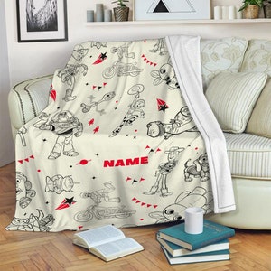 Peut inclure: Un plaid en polaire couleur cr&egrave;me orn&eacute; d'un motif r&eacute;p&eacute;titif en traits noirs repr&eacute;sentant divers personnages anim&eacute;s, dont un ranger de l'espace, un cow-boy et une fourchette-jouet. Le design inclut des accents rouges, des &eacute;toiles et le mot NAME.