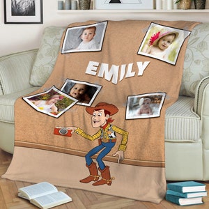 May include: A brown and tan fleece blanket with a corkboard design. The blanket features a cartoon image of Woody from Toy Story holding a camera. There are several photos of babies on the blanket, along with the name "EMILY".